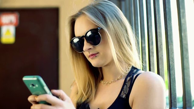 Punk girl wearing sunglasses and texting on smartphone next to the gate
