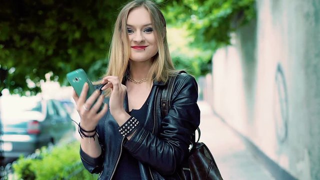 Punk girl texting on smartphone while standing on pathway and smiling to the camera
