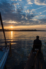 Mężczyzna wędkujący na pomoście nad jeziorem koło zacumowanego jachtu. W tle zachód słońca.