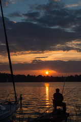 Mężczyzna wędkujący przy zachodzącym słońcu obok zacumowanego jachtu.