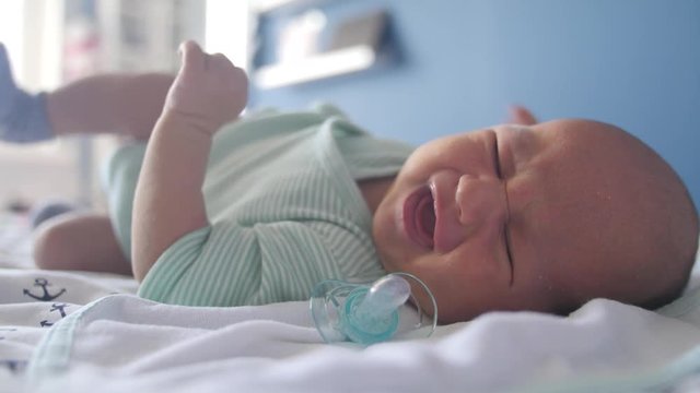 Caucasian newborn lying in a bed, letting his baby dummy to fall out and crying
