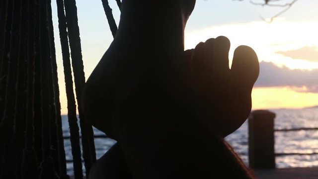Close up of feet stretching and flexing in a seaside hammock at sunset; shot then reframes to a medium CU. (POV of person lying in the hammock). Sea is in the nearby backgroun