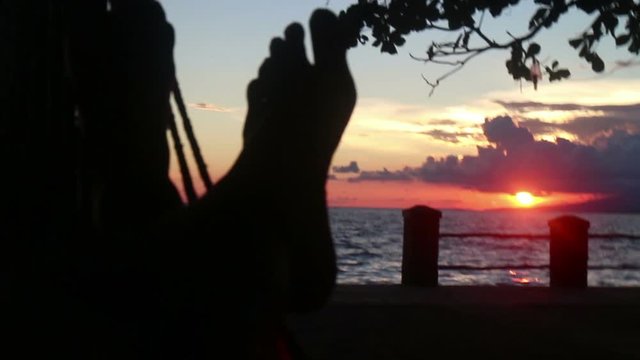 Medium point of view of person's feet in silhouette in a hammock at dusk with deep red sunset by the sea. (POV of person lying in the hammock). Sea is in the nearby background