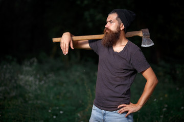 Brutal brunette bearded man in warm hat with a hatchet in the woods on a background of trees