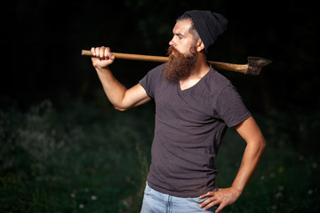 Brutal brunette bearded man in warm hat with a hatchet in the woods on a background of trees