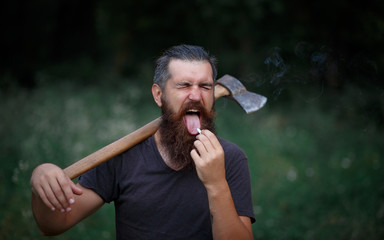 Fototapeta premium Brutal brunette bearded man in warm hat with a hatchet in the woods on a background of trees