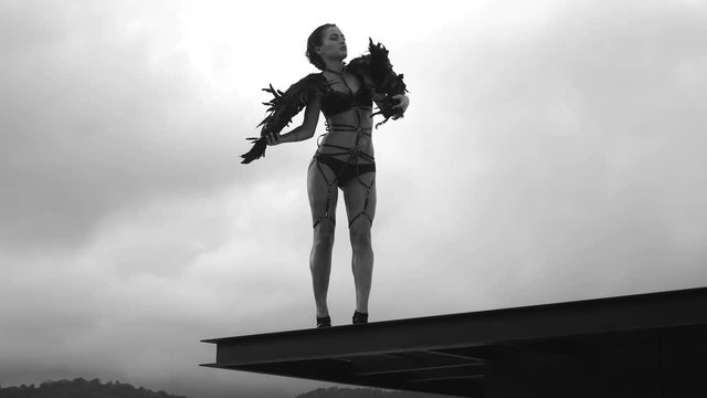 Black and white video of beautiful seductive angel woman wearing lingerie and leather belts standing on the roof with wind in her wings over cloudy sky