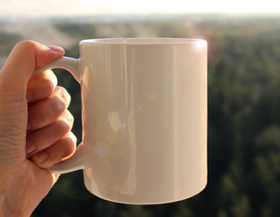 White mug in hand, Mockup. Misty autumn forest on the background.