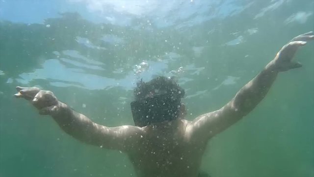 Wide shot of man wearing VR headset descends deeper under the ocean, with arms spread.  He slowly 'looks around', suggesting an immersive virtual experience (60 fps)