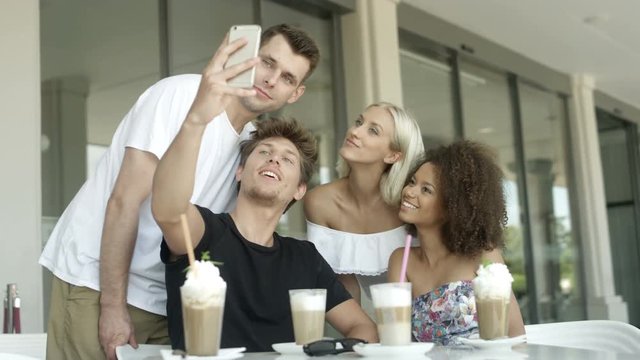 Group of young people sitting in a restaurant and taking a selfies. Group of friends making selfie photos. Young multi-ethnic people taking pictures by smartphone in outdoor restaurant.