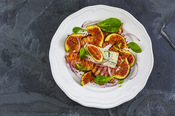 Figs and gorgonzola and fried bacon with small lettuce leaves served on a white plate. 
