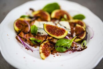 Figs and gorgonzola and fried bacon with small lettuce leaves served on a white plate. 
