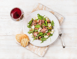 Prosciutto, arugula, basil, figs and mozzarella salad served with slices of bread on rustic board with glass of red wine over white wooden background, top view