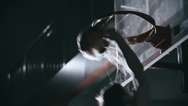 Slow motion shot of basketball player shooting the ball directly through the basket with one hand in the darkness and making wet net splashing water droplets