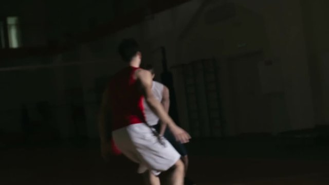 Basketball player dribbling a ball and scoring a point while playing against opponent at dark court in slow motion