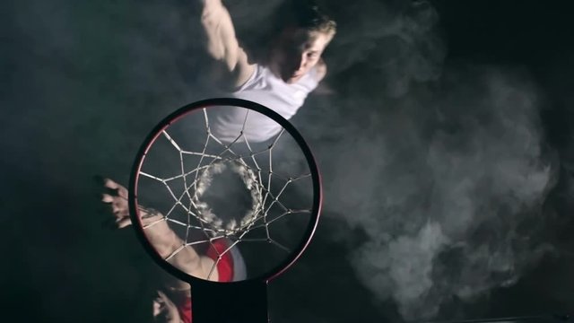 Top view of two basketball opponents jumping in the air and one man slamming ball in the net in the dark court with smoke in slow motion
