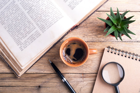 Cup of coffee, old open book, succulent, pen, notebook and magnifier on rustic wooden background. Office desk table. Top view
