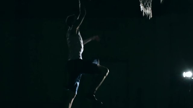 Side view of basketball player jumping and shooting ball into the net with one hand in slow motion in the darkness