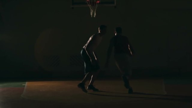 Basketball player dribbling a ball, running and throwing it into the net while his opponent trying to stop him in dark court