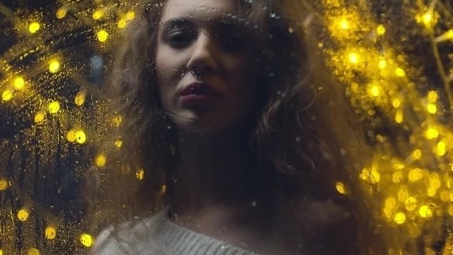 Low angle shot of curly woman standing behind foggy window in dark studio decorated with yellow fairy lights, then wiping it with hand and looking at camera