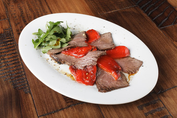 Baked roast beef with rucola and bell pepper in a white oval plate. Wooden background.