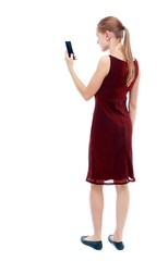 Naklejka premium back view of standing young beautiful woman using a mobile phone. girl watching. Isolated over white background. The girl in the maroon sleeveless dress looks into the smartphone.