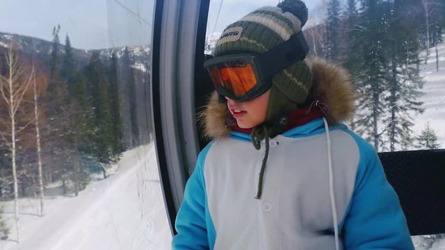 Happy boy in sunglass mask, rises to the top in the cabin cable car at the ski resort on the background of mountains in slowmotion. 1920x1080