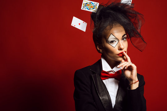 Portrait of a young beautiful lady croupier with an artistic make up joker and flying cards around on the red background. Gamble and casino concept. Studio shot. Copy-space