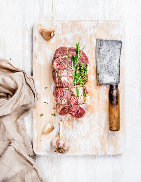 Raw uncooked roastbeef meat cut with rosemary, thyme and garlic and butcher knife on old white painted wooden background, top view, vertical composition