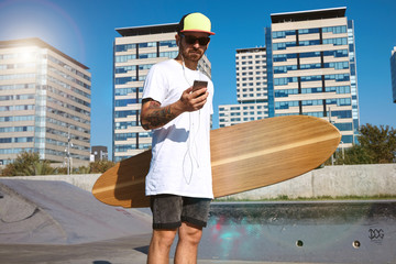 Skater in sunglasses and baseball cap looking at his smartphone and listening music through earplugs headphones, wearing blank white t-shirt and holding big wooden longboard in tattooed hand