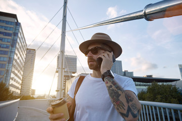Portrait of a young bearded and tattooed traveler in fedora hat and white unlabeled t-shirt talking...