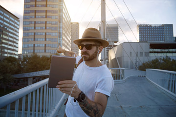 Bearded and tattooed hipster on a city bridge showing an office building to his friend during a video call at sunset