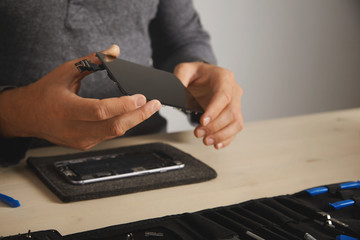 Master holds new screen for replacement above disassembled smartphone in his laboratory, tool kit with instruments in front of him on white table, space for your text on right