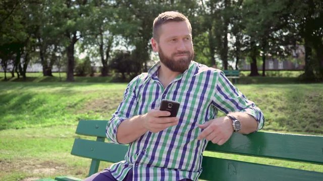 Man browsing smartphone. He is sitting on bench in the park. He is dressed in blue shorts and checkered shirt. He has beard. Steadicam.
