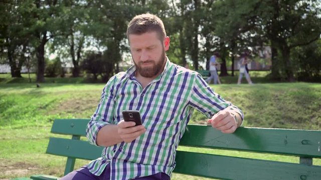 Man browsing smartphone smiles for camera. He is sitting on bench in the park. He is dressed in blue shorts and checkered shirt. He has beard. Steadicam.

