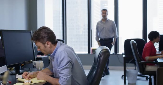 Bored business man leaves desk to interrupt co-worker and engage in conversation.  Wide shot, recorded in real time in open-plan city office.