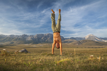 Shirtless man doing a handstand in a green field. Wildlife landscape, mountains background. Freedom and naturalness icon