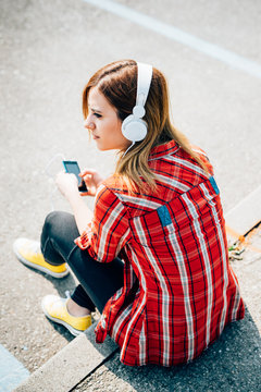Back view of young beautiful caucasian blonde hair woman listening music with headphones and smart phone hand hold outdoor in the city - music, technology, communication concept