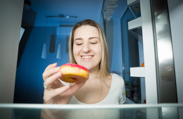 Portrait of smiling woman taking donut from refrigerator shelf a