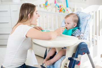 Beautiful woman feeding her baby boy in highchair at living room