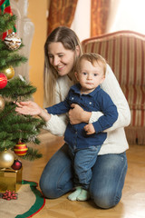 Smiling woman decorating Christmas tree with her 10 months old b