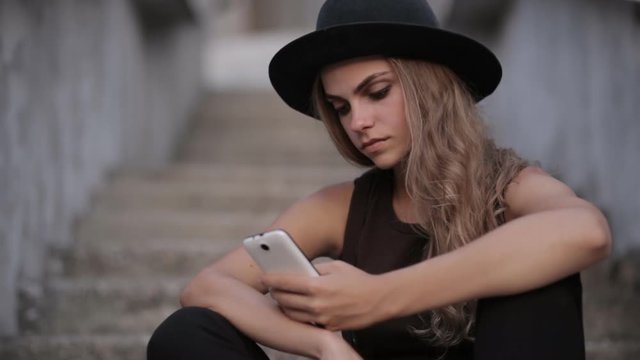 pretty young girl in a black hat is typing a message on a smartphone