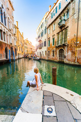 Woman sits near a canal and admires gondolas and architecture of Venice. Italy.