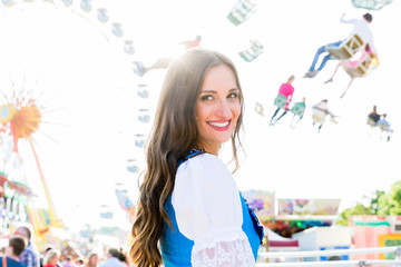 Frau im Dirndl steht lächelnd vor Riesenrad und dem Kettenkarussell auf bayrischem Volksfest