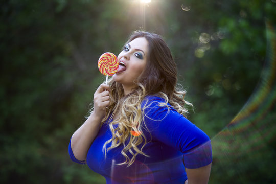 Young beautiful caucasian plus size model in blue dress outdoors, xxl woman on nature licking a lollipop