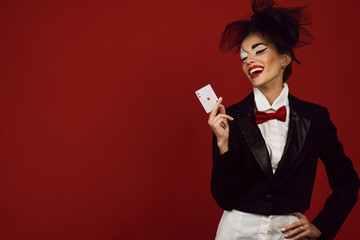 Portrait of a young beautiful lady croupier with an artistic make up joker on the red background holding an ace card and laughing. Gamble and casino concept. Winner. Studio shot. Copy-space