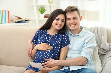 Beautiful young pregnant couple with ultrasound image sitting on sofa