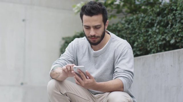 Young Man Sitting on the Steps Outdoors and Using Mobile Phone. Modern Urban Environment. Shot on RED Cinema Camera in 4K (UHD).