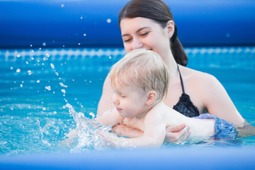 Fototapeta na wymiar Happy young mother and her one year-old son, adorable laughing baby boy having fun together in an outdoor swimming pool on a hot summer day during vacation