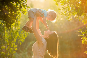 Happy young mother playing with her littlte baby son on sunshine warm autumn or summer day. Beautiful sunset light in the apple garden or in the park. Happy family concept